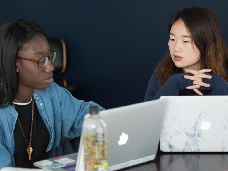 Two female students talking while working at laptops.